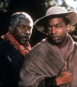 Older Black man in gray hair standing next to a younger black man draped in a blanket and wearing a hat. 