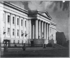 White building with columns in the front. Walkers are in front of the structure in the historic photograph.